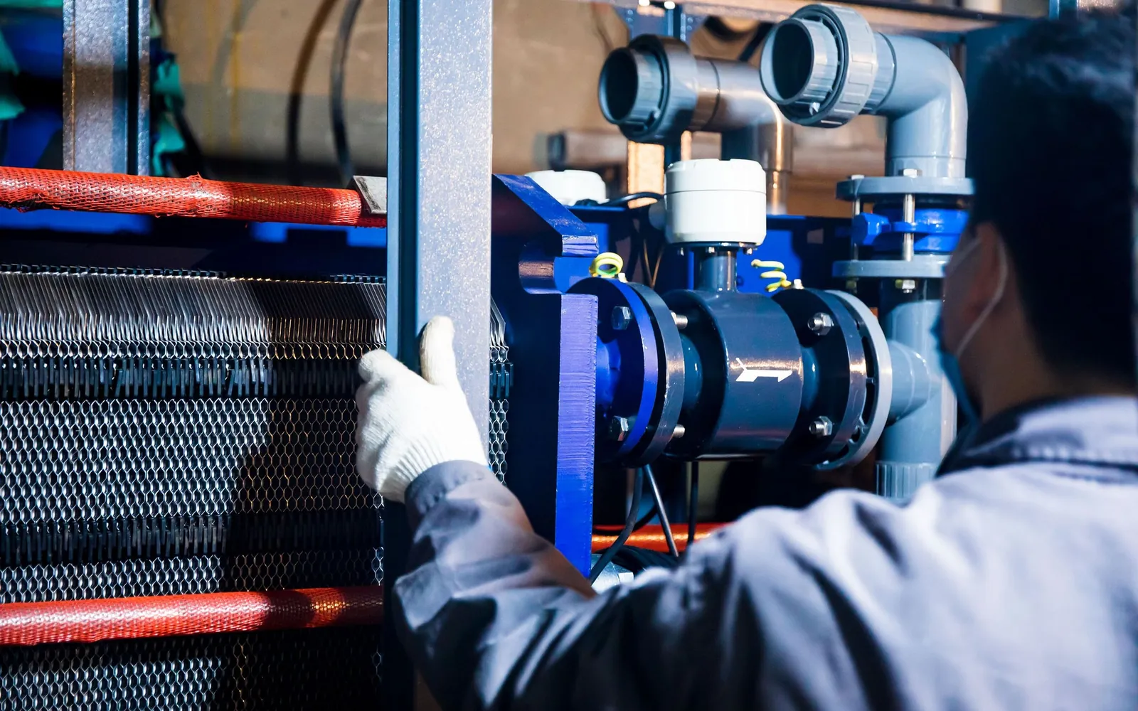 Gloved technician inspecting piping and heat-transfer equipment in an industrial work area.