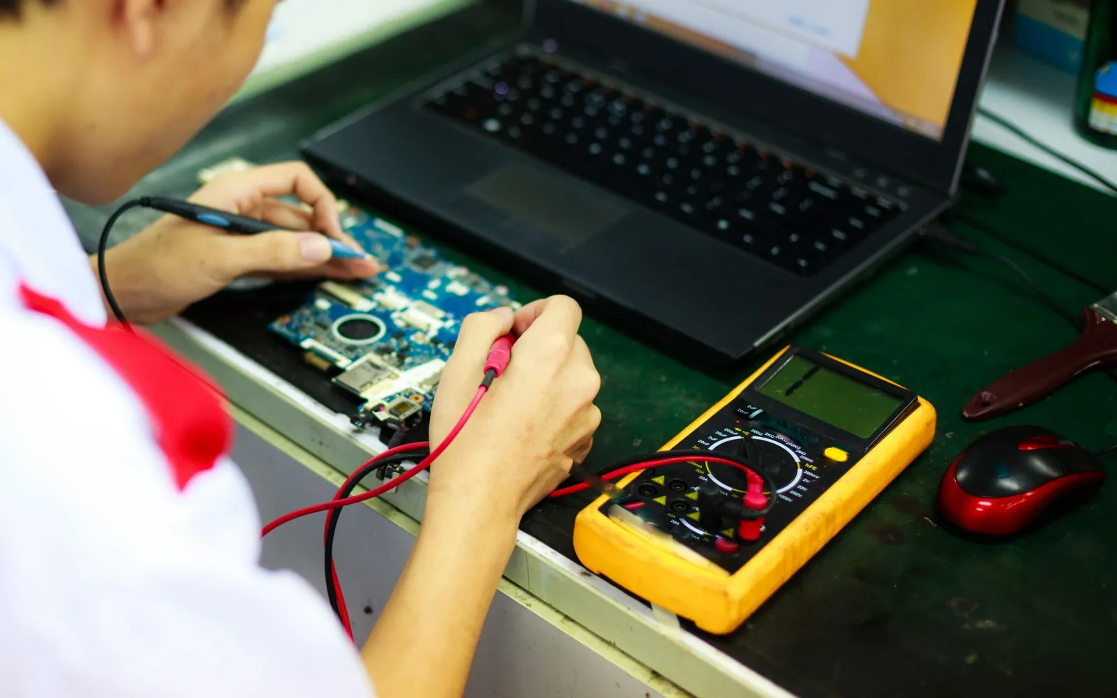 Adult technician using a meter and laptop while practicing electronics repair at a workbench.