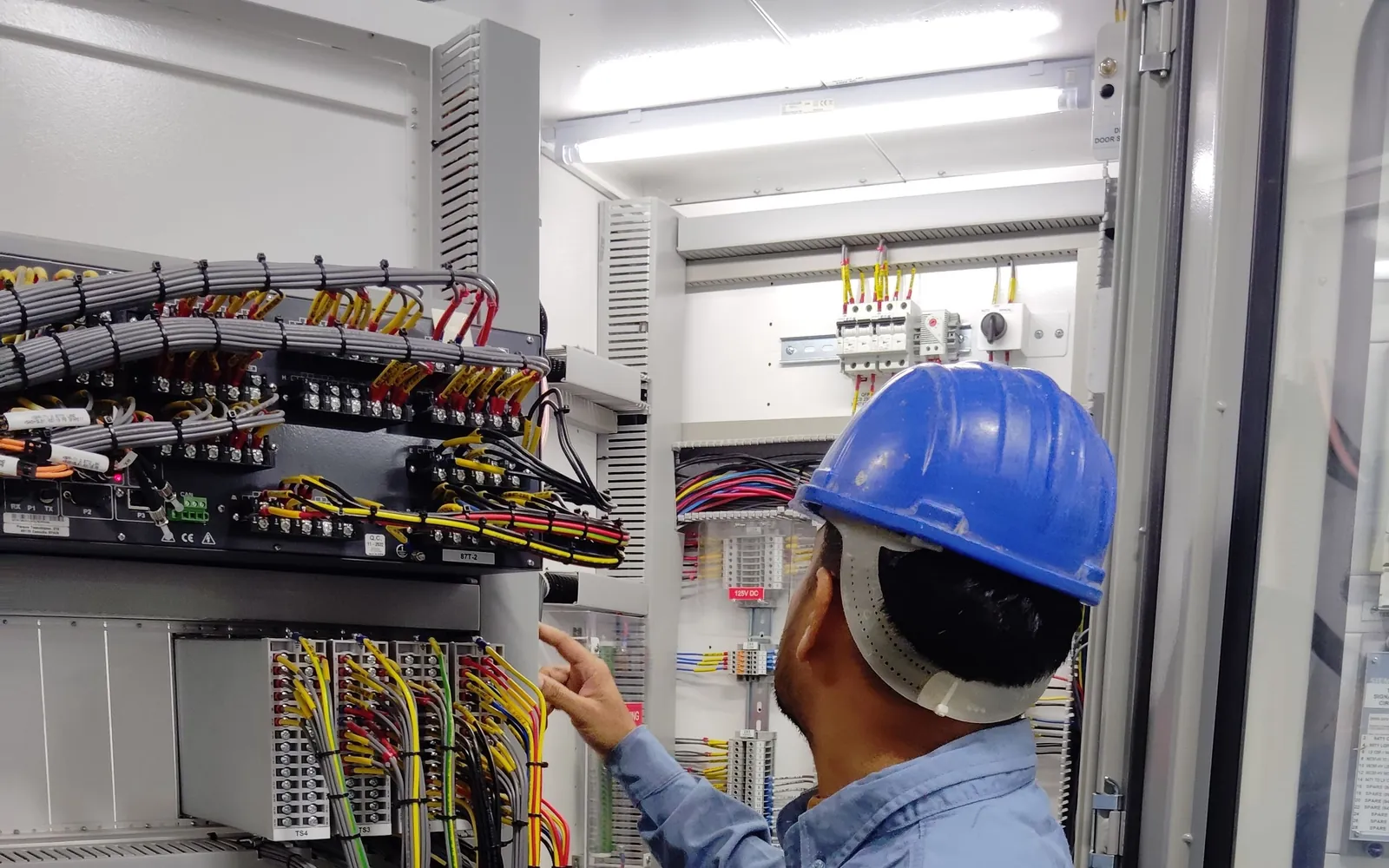 Hard-hat technician inspecting wiring inside an open industrial control cabinet.