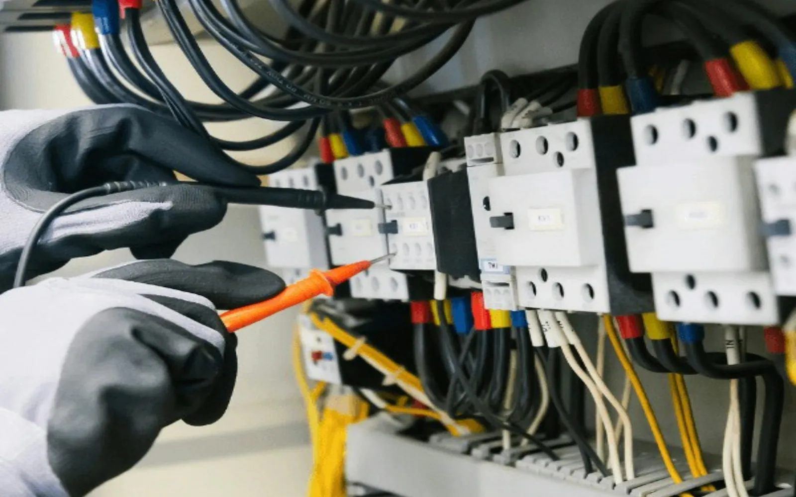Gloved technician using insulated test probes inside an industrial electrical control panel.